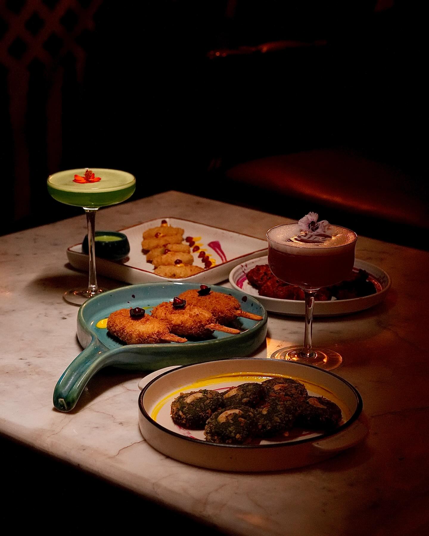 A spread of cocktails, tandoori and small plates on a dark table