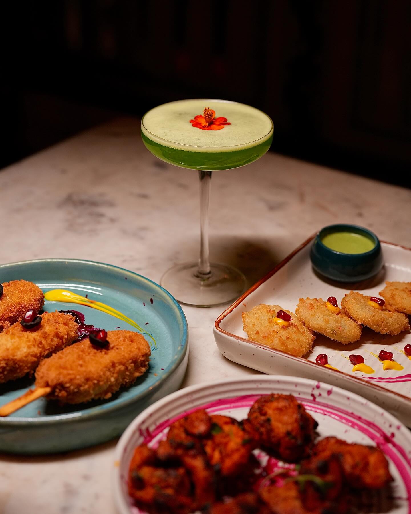A green coupe cocktail beside fried plates and bright chutneys